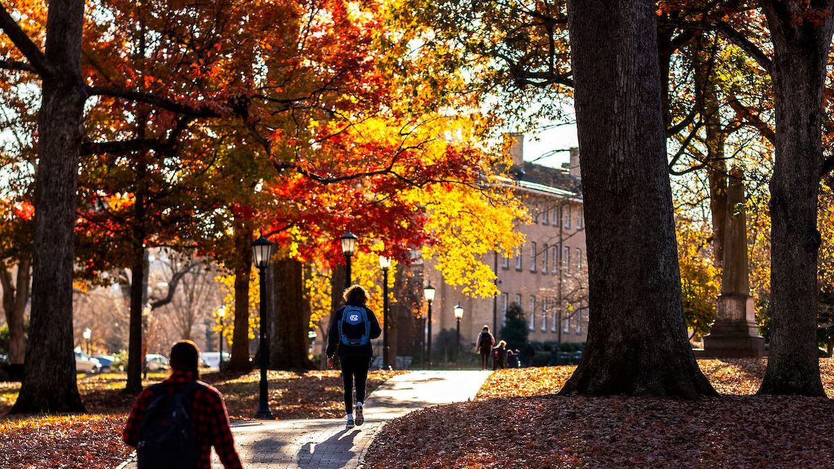 Students walk across pathways on the campus of UNC-Chapel Hill underneath trees with fall foliage