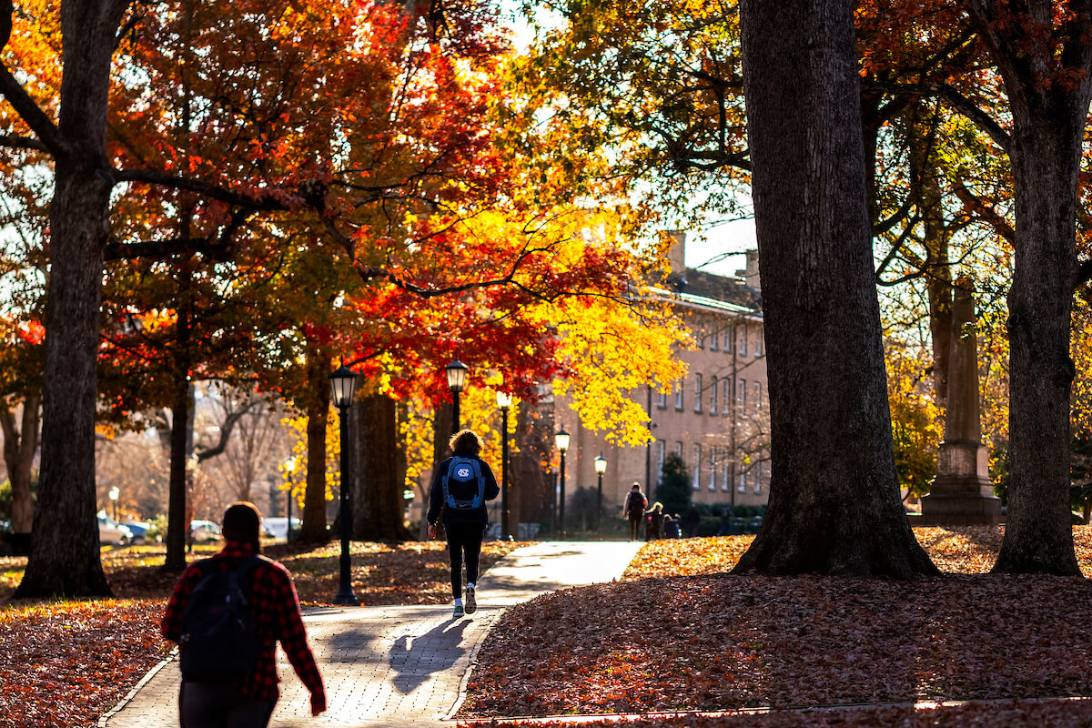 Students walk across pathways on the campus of UNC-Chapel Hill underneath trees with fall foliage