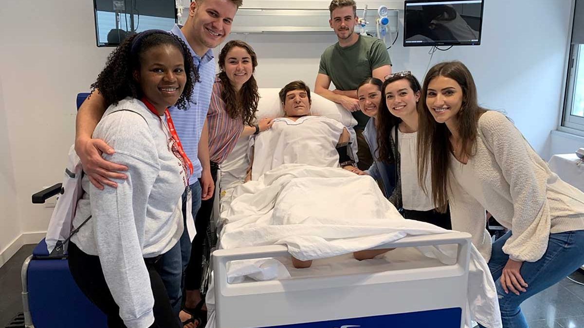 A group of Carolina nursing students pose around a hospital bed in a simulation exercise.