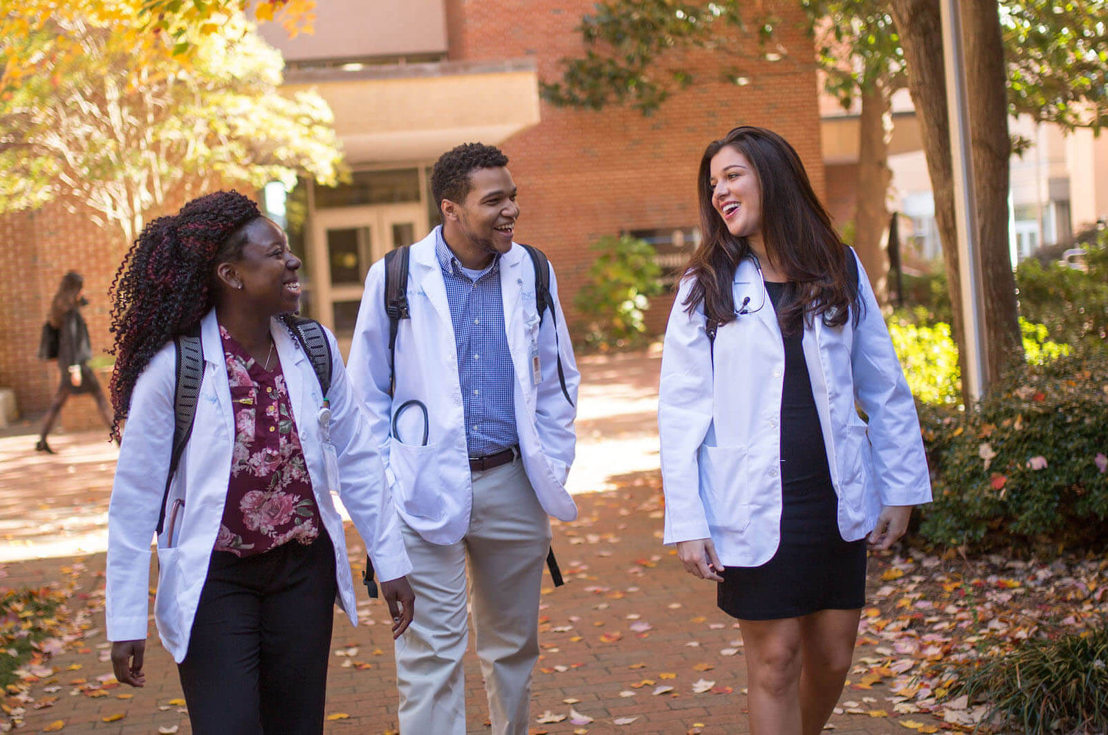 3 students in white lab coats walking on the campus of UNC.