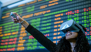 Carolina student wearing virtual reality goggles in front of a stock ticker board