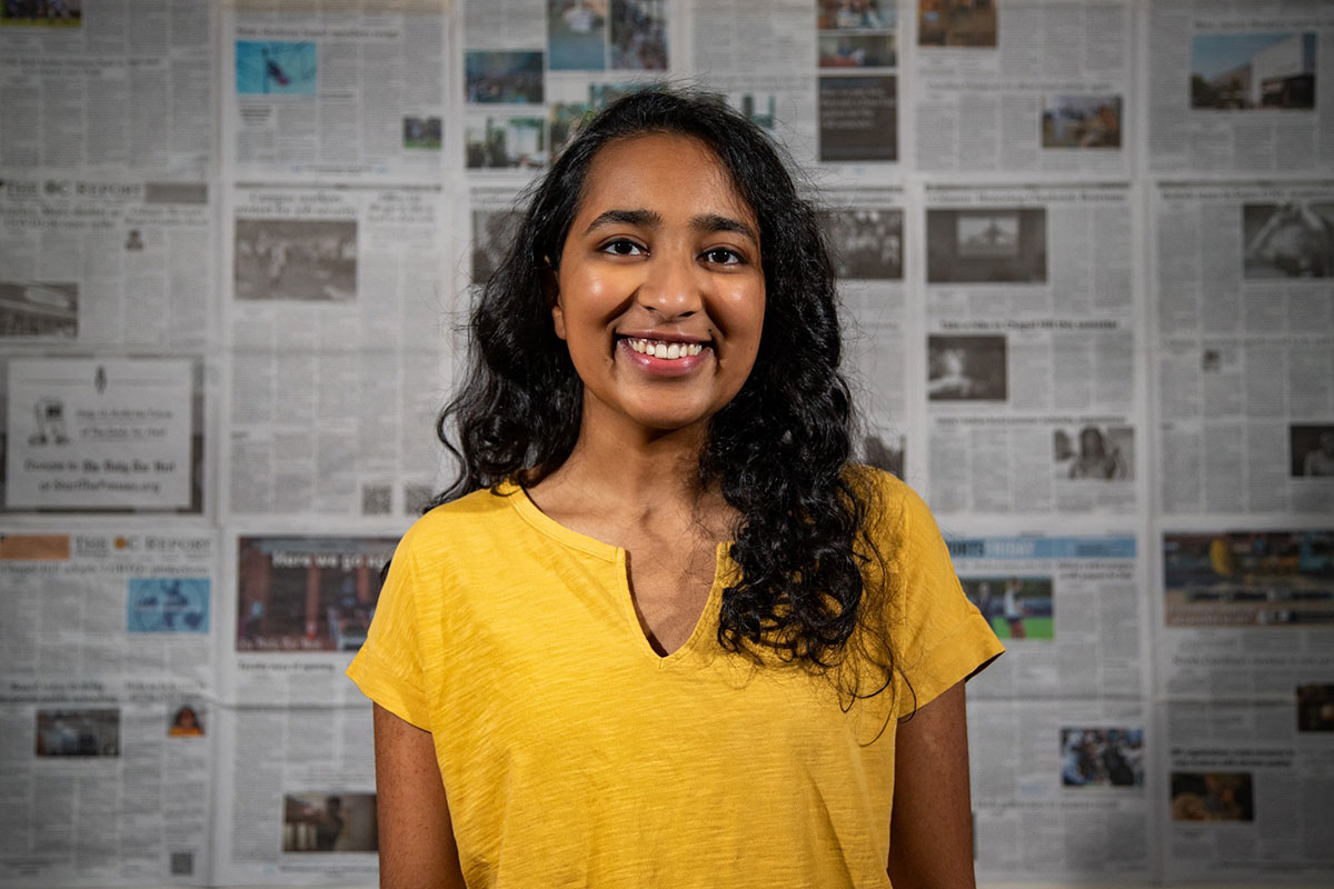 Pravenna standing in front of a wall covered in newspaper articles, smiling
