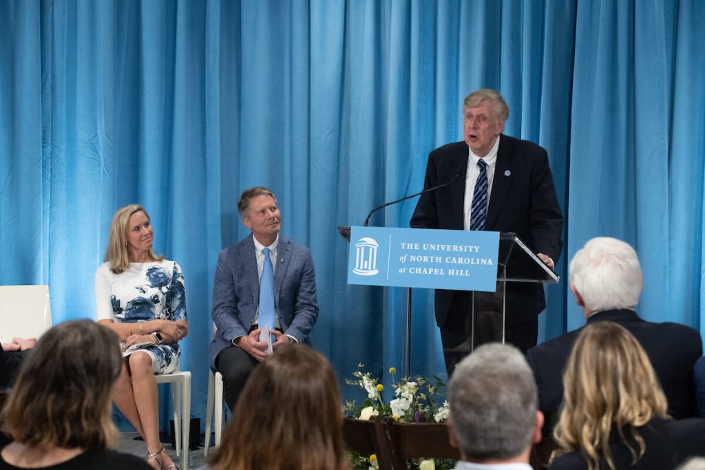 Don Curtis speaks at a lectern at the opening celebration of the Curtis Media Center, with Chancellor Kevin Guskiewicz and other dignitaries seated beside him
