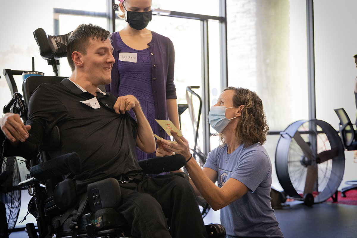 Kara Hume kneels beside a young man in a wheelchair during her adaptive fitness class
