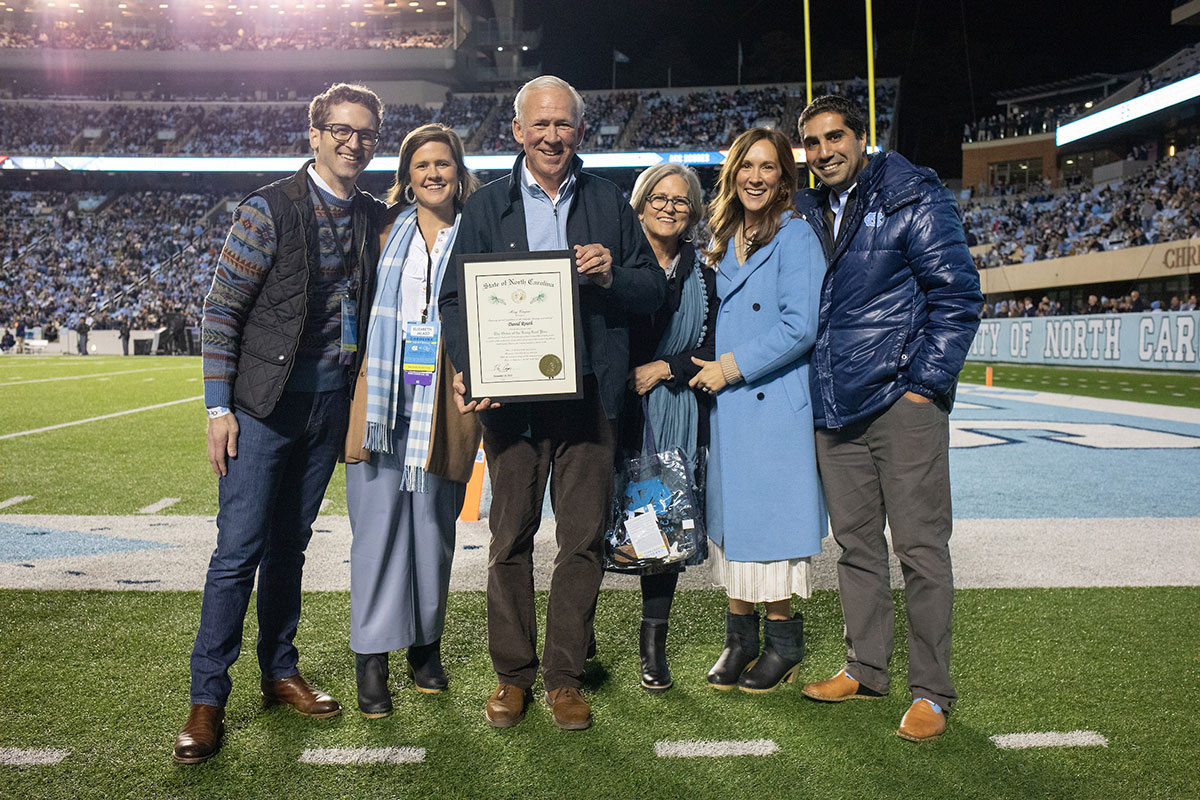 David Routh stands in the center of his family holding the Order of the Long Leaf Pine Award with Eddie Smith Field in the background.