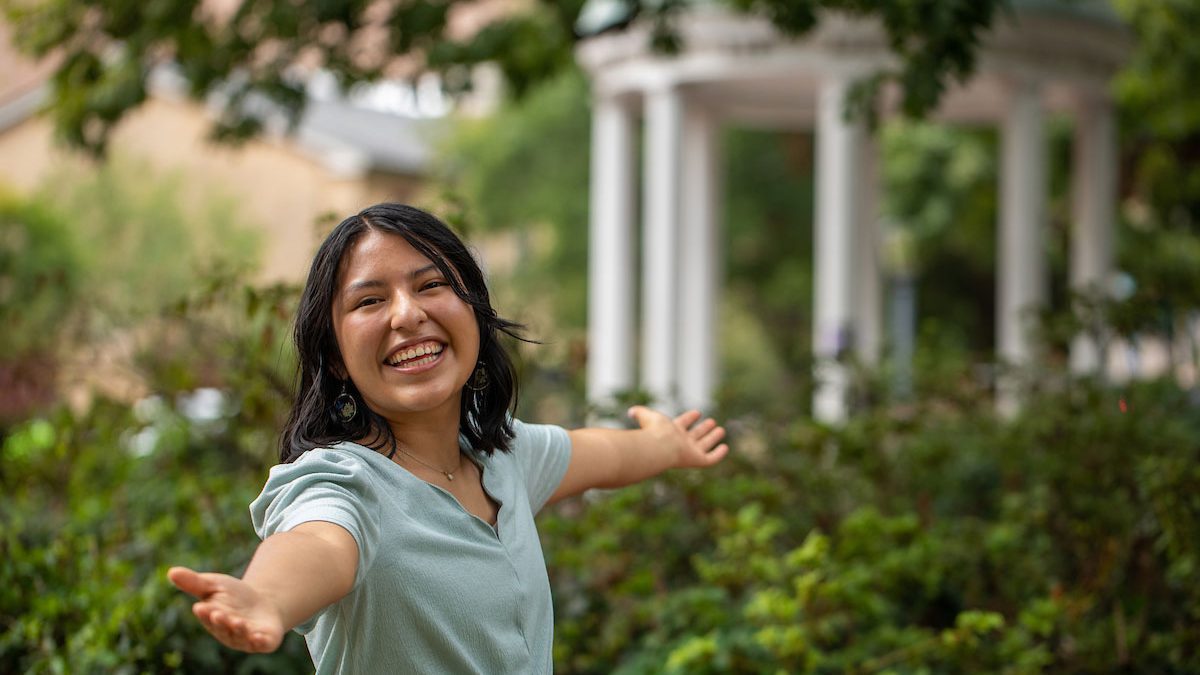 Brenda Palacios Rodriguez stands in the foreground with arms spread wide and a huge smile on her face. The Old Well at UNC-Chapel Hill is in the background.