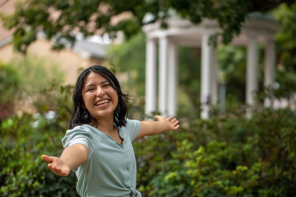 Brenda Palacios Rodriguez stands in the foreground with arms spread wide and a huge smile on her face. The Old Well at UNC-Chapel Hill is in the background.
