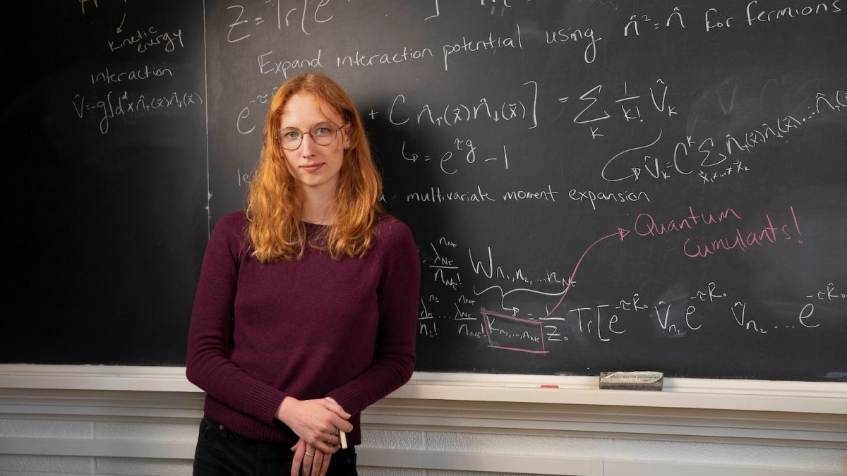 Doctoral student Kaitlyn Morrell poses in front of a blackboard that shows physics equations.