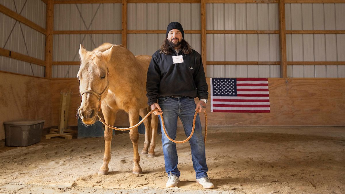 Veteran Matthew Colon stands in a barn with a horse, facing the camera. An American flag is in the background.